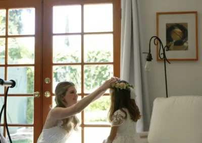 A woman in a white dress adjusts a young girl's hair near French doors in a bright room.