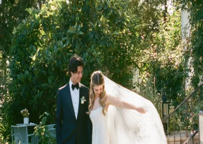 A bride and groom walking together outdoors, surrounded by greenery.