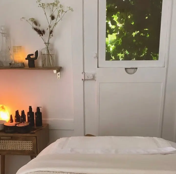 Massage room with a lit salt lamp, essential oil bottles, and a window view of green foliage.